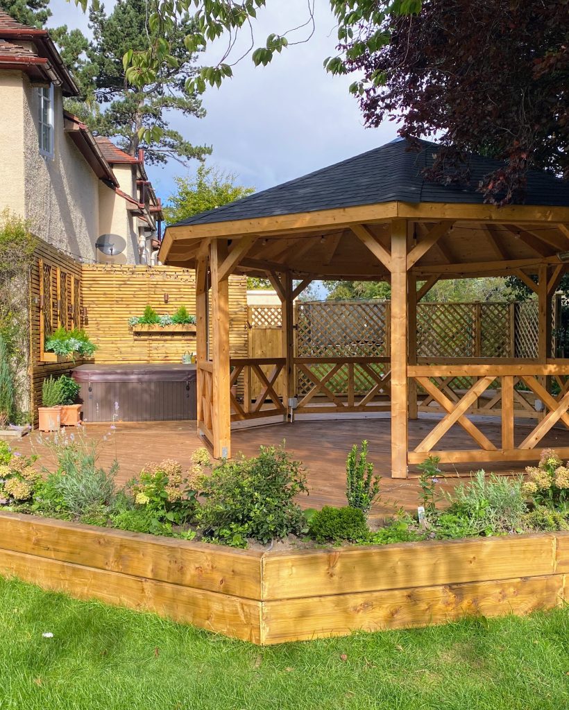 Wooden gazebo in a landscaped garden with plants and a hot tub.