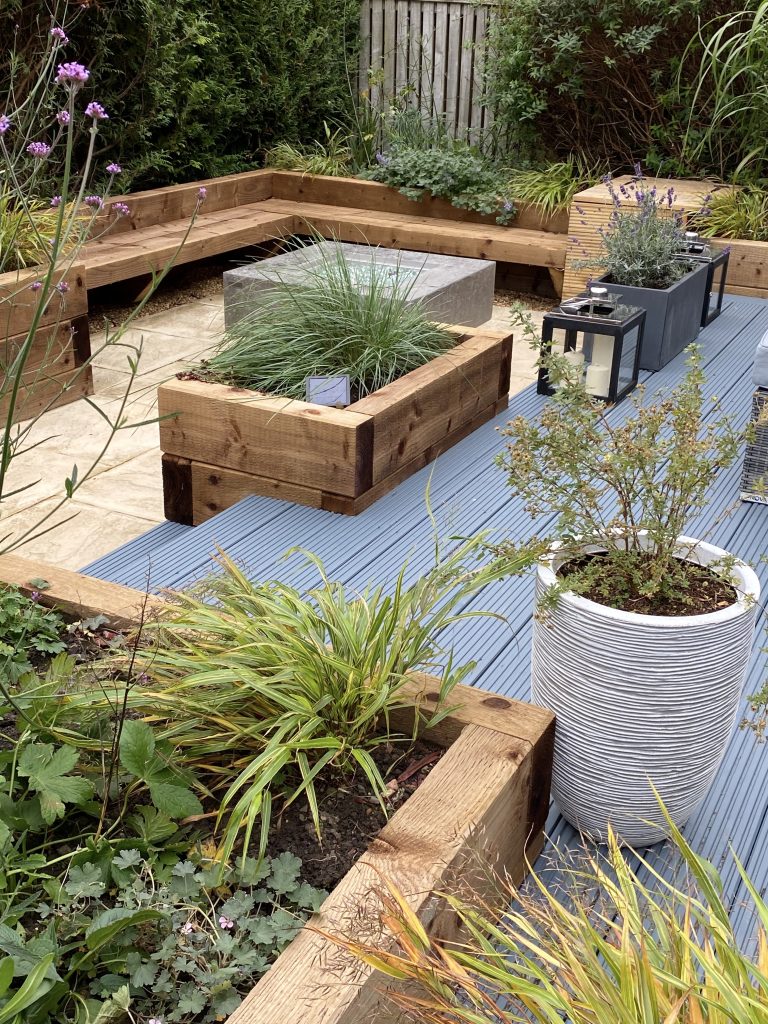 Raised wooden planters with modern blue decking and a firepit centerpiece